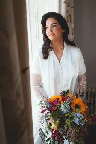 Bride in a robe holding a sunflower bouquet in soft window light during morning preparations at Millbrook Lodge
