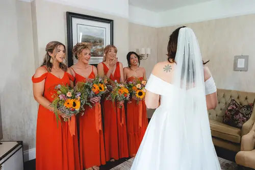 Bride laughing with bridesmaids in orange dresses, holding sunflower bouquets during spring wedding prep at Millbrook Lodge