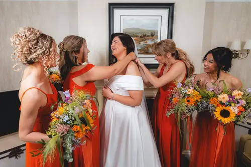 Bridesmaids in orange dresses fastening the bride’s gown during morning preparations at Millbrook Lodge