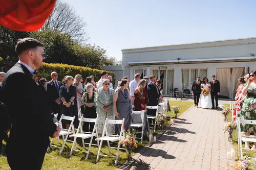 Bride walking down the outdoor aisle at Millbrook Lodge, with guests standing in the spring sunshine