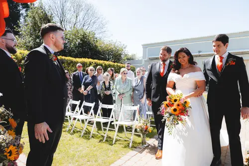 Bride walking down the outdoor aisle at Millbrook Lodge as the groom waits in sunny spring light
