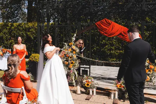 A couple laugh during their sunny outdoor ceremony at Millbrook Lodge, Ballynahinch, with bridesmaids and floral decor nearby