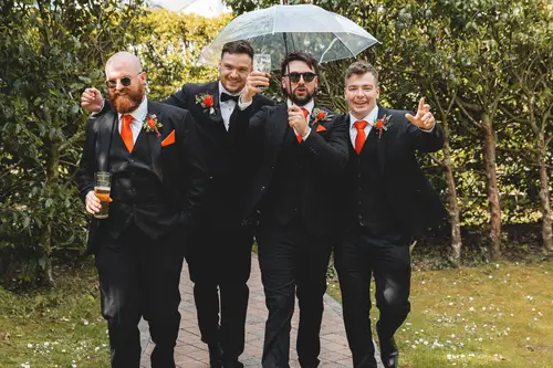 Groomsmen in suits with orange ties laughing under an umbrella on the garden path at Millbrook Lodge