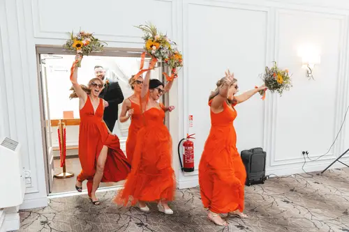 Bridesmaids in orange dresses carry bouquets into the bright reception at Millbrook Lodge, Ballynahinch