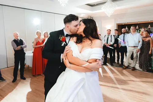 Couple kiss during their first dance at Millbrook Lodge, with guests gathered around the warmly lit dance floor