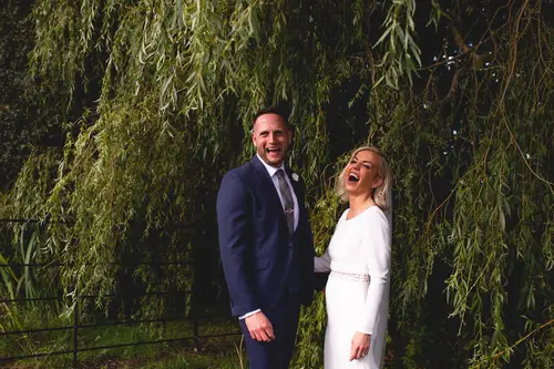 Bride and groom laughing together under willow trees at Old Rectory in Killyman, surrounded by soft greenery and natural light.