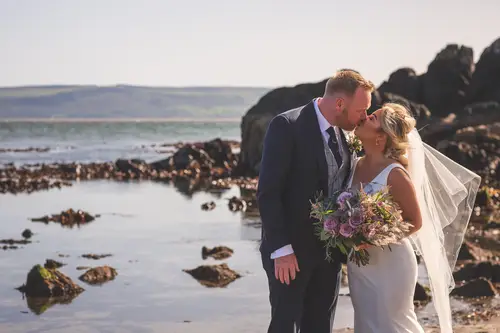 Bride and groom kiss on coastal rocks at golden hour near Redcastle Hotel, Moville, with bouquet and veil in soft seaside light