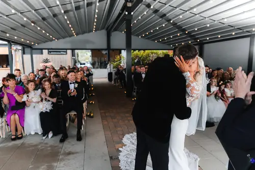 First kiss during a ceremony on the covered terrace at Rossharbour Resort, with guests applauding under string lights
