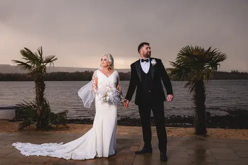 Bride and groom holding hands by Lough Erne at Rossharbour Resort, with sunset light and a dramatic sky