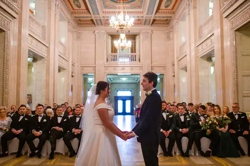 Bride and groom holding hands during their ceremony at Stormont in Belfast, surrounded by guests in soft indoor light.