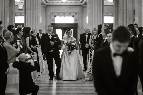 Black and white photo of a bride walking down the aisle at Stormont in Belfast during an indoor wedding ceremony
