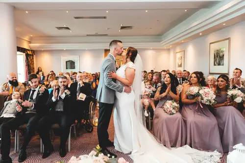 First kiss during an indoor wedding ceremony at Stormont, with guests and bridal party applauding in soft light.