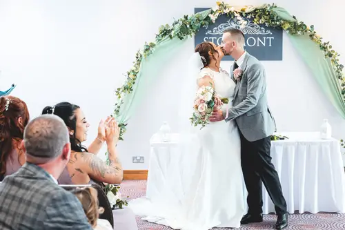First kiss during an indoor wedding ceremony at Stormont, Belfast, with guests applauding beneath a floral arch