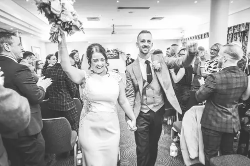 Black and white aisle exit at Stormont in Belfast, with the couple smiling as guests cheer and the bouquet is raised.