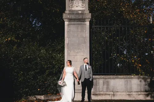 Bride and groom holding hands by a sunlit stone gate at Stormont in Belfast on an autumn wedding day