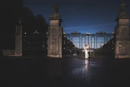 Bride and groom in a backlit night portrait by the gates at Stormont, Belfast, with rain on the pavement.