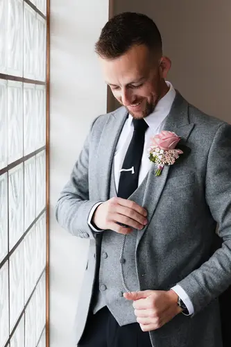 Smiling groom in a grey suit with buttonhole by window light during morning prep at Stormont, Belfast