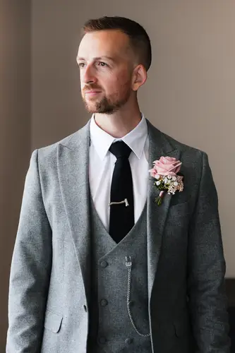 Groom in a grey suit adjusting his buttonhole by window light during the wedding morning at Stormont, Belfast