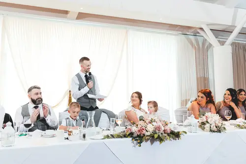 Joyful wedding speeches at Stormont in Belfast, with the bride, groom and bridal party laughing at the flower-filled top table