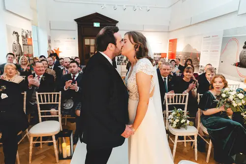 First kiss during an Ulster Museum wedding ceremony in Belfast, with guests applauding in soft natural light