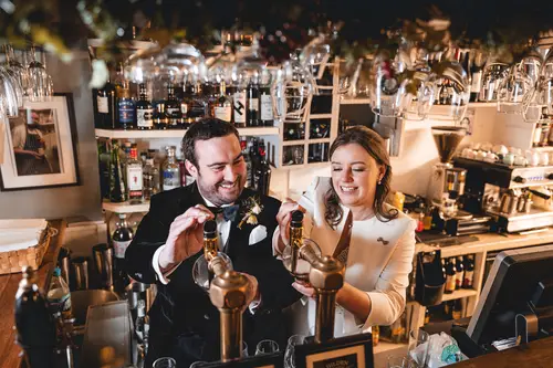 Bride and groom pouring drinks at the Ulster Museum bar in Belfast, captured in warm light during their wedding reception