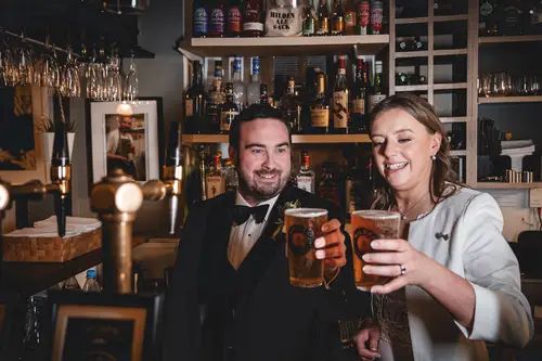 Bride and groom sharing a relaxed drinks toast at the Ulster Museum bar in Belfast, captured in warm indoor light