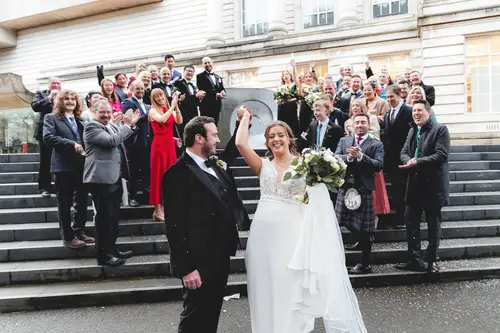 Bride, groom and guests sharing a high five on the steps of Ulster Museum in Belfast during a joyful confetti moment