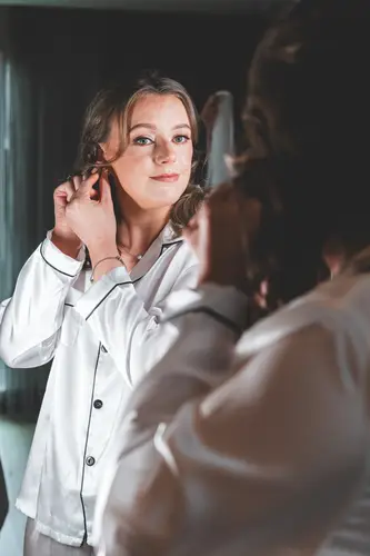 Bride in pyjamas putting on earrings in a mirror during calm morning prep at Ulster Museum, Belfast